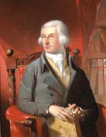 White man wearing wig, grey coat, yellow vest, and white cravat, seated in red armchair next to bookshelf and holding a book.